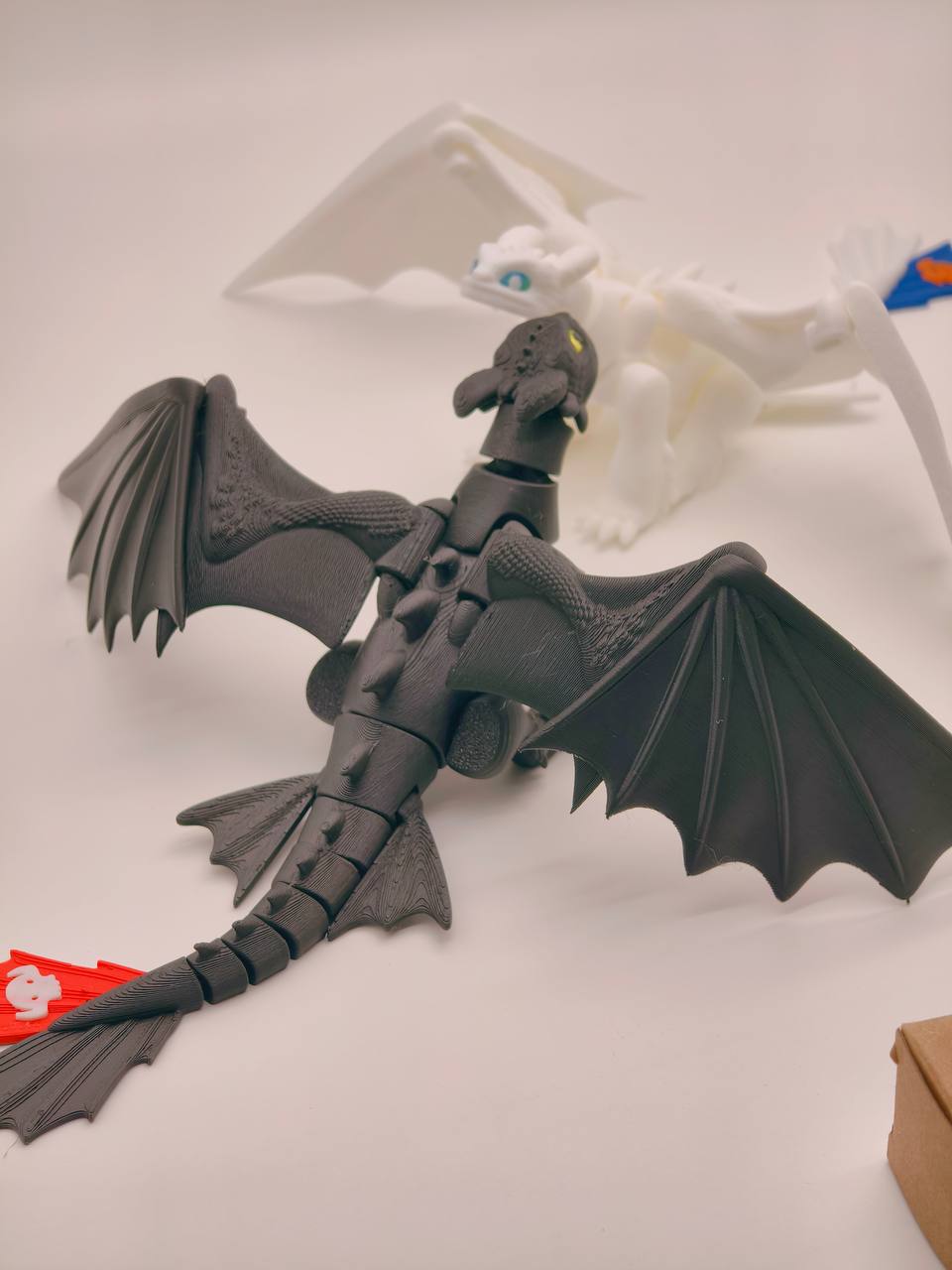 A black articulated dragon toy with spread wings, shown on a light gray surface with a white dragon figure blurred in the background.